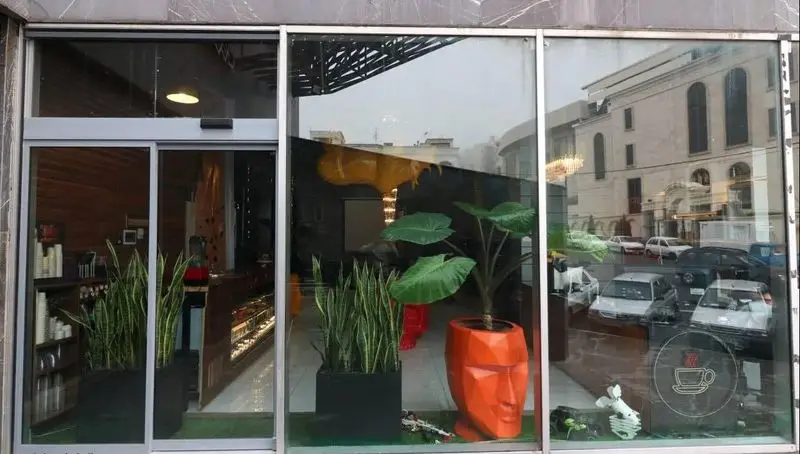 ویترین کافه رستوران پلاس شیم، نمایشگر فضای دلنشین با گیاهان سبز و پیشخوان، لوگوی قهوه روی شیشه و انعکاس محیط شهری.