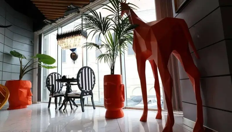 دکوراسیون داخلی مدرن کافه رستوران پلاس شیم با پنجره‌های بزرگ، گیاهان شیک، صندلی‌های راه‌راه و مجسمه زرافه نارنجی.