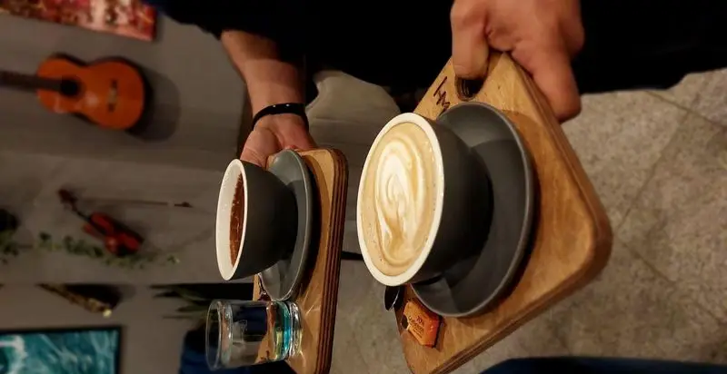 سرو دو فنجان قهوه (یکی با لاته آرت زیبا) در سینی چوبی توسط پرسنل کافه و رستوران تراس لانژ؛ فضایی دلنشین با دکوراسیون موسیقی.