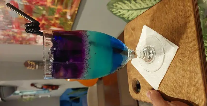 یک نوشیدنی خنک و رنگارنگ لایه‌ای با دانه‌های شناور و نی، سرو شده در لیوانی بلند روی سینی چوبی در کافه و رستوران تراس لانژ.