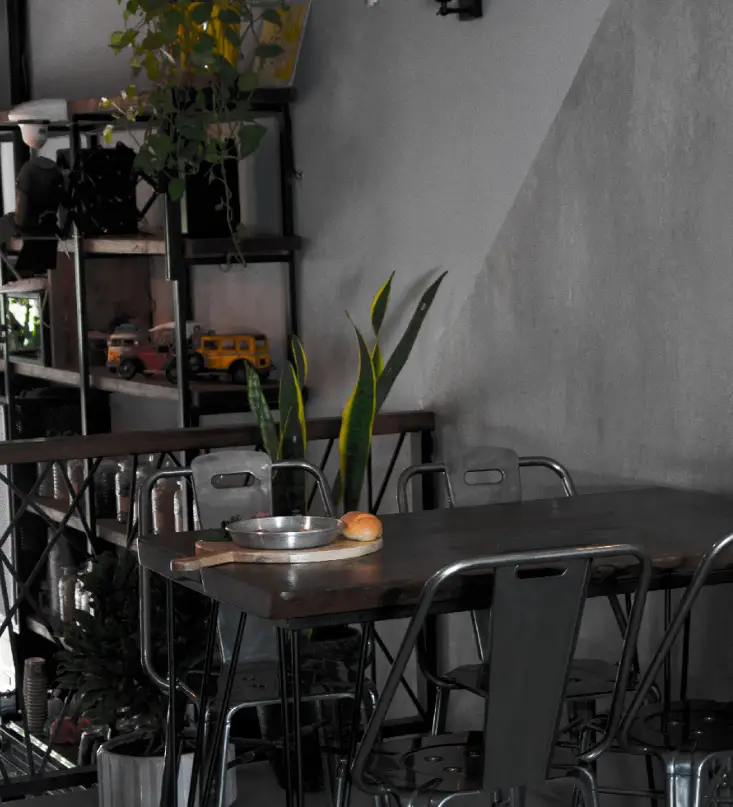 فضای داخلی کافه ۰۸: میز چوبی با صبحانه (پن و نان)، صندلی‌های صنعتی فلزی، دکوراسیون خاص با گیاهان و ماشین‌های مینیاتوری. محیطی دنج و مدرن.