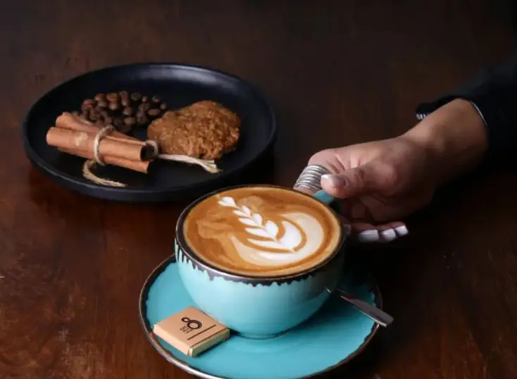 لاته چشم‌نواز کافه ۰۸ با لته آرت هنرمندانه، فنجان فیروزه‌ای زیبا، در کنار کوکی، دارچین، دانه قهوه و شکلات. تجربه دلنشین در کافه ۰۸.