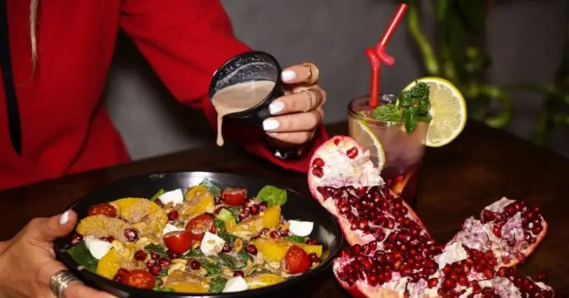 صحنه لذت‌بخش از مشتری در کافه ۰۸ که سس مخصوص را روی سالاد میوه و سبزیجات تازه شامل انار و پرتقال می‌ریزد، در کنار یک نوشیدنی.