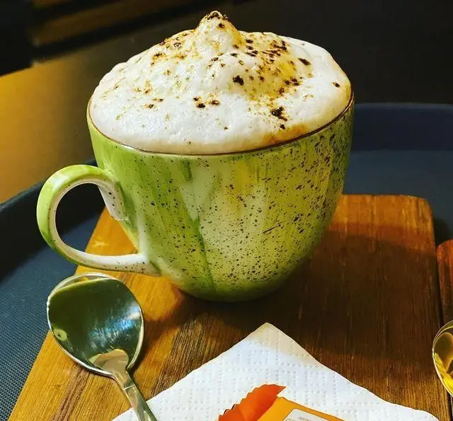 یک فنجان بزرگ کاپوچینو لذیذ کافه برلین با فوم شیر غلیظ و پودر قهوه روی آن، سرو شده در فنجان سبز هنری. تجربه‌ای دلپذیر از بهترین نوشیدنی‌های گرم در فضایی آرام.