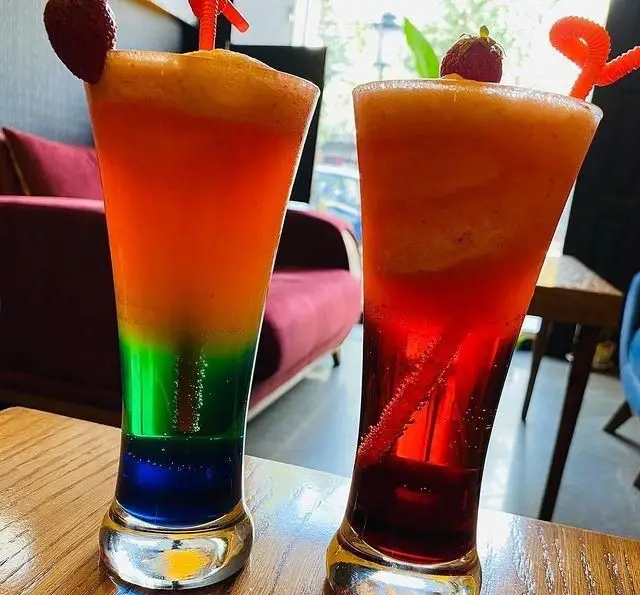 دو نوشیدنی لایه‌لایه رنگارنگ کافه برلین: اسموتی رنگین‌کمان و کوکتل قرمز-نارنجی. با توت فرنگی و نی، طراوت‌بخش روزهای گرم.