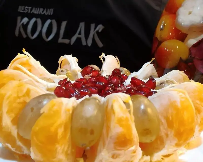 دسر میوه جذاب و تازه در کافه قلیان و سفره خانه سنتی کولاک: پرتقال برش خورده با دانه های انار و انگور، لوگوی رستوران در پس زمینه.