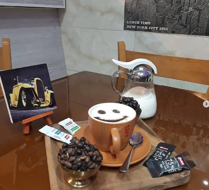 تصویر فنجان قهوه لاته با لبخند در کافه آرمان، همراه با دانه های قهوه، شکلات تلخ و شکر. فضایی آرام و دلپذیر.