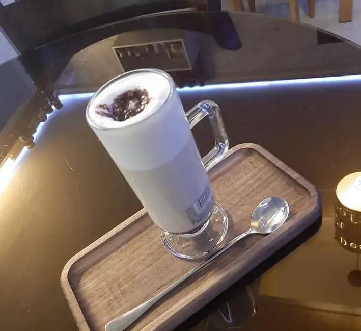 نوشیدنی لاته یا شیر قهوه فومی و جذاب کافه آرمان در لیوان دسته دار شیشه‌ای روی سینی چوبی، با قاشق بلند و میز براق کافه.