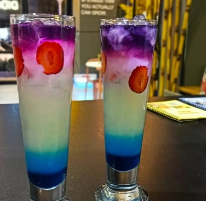 دو لیوان موکتل رنگین‌کمان جذاب و خنک با لایه‌های میوه‌ای بنفش، صورتی، آبی و توت‌فرنگی تازه، از نوشیدنی‌های تابستانی کافه و فست فود دیدومک.