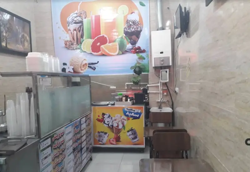 تصویر داخلی مغازه ویتامینه سعید با پیشخوان، دستگاه آبمیوه، بستنی، دسر، پوستر میوه و نوشیدنی‌های تازه و میز و صندلی برای مشتریان.