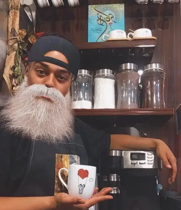 تصویری جذاب از باریستای برگر دلشاد با ریش سفید، فنجان قهوه ای با طرح قلب در دست. لذت نوشیدنی های خاص در کافه ما.