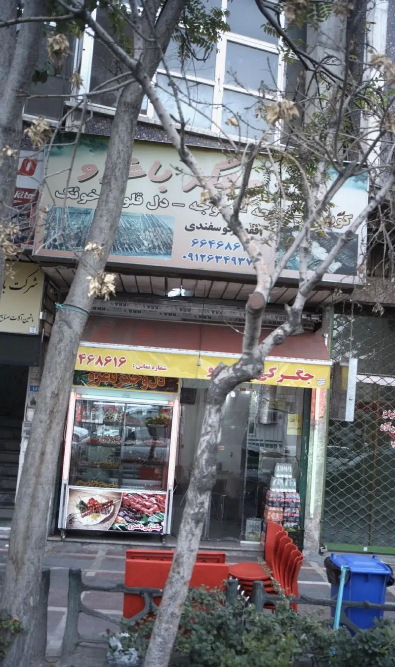 نمای خارجی جگرکی باغرو با تابلوهای تبلیغاتی کباب دل، جگر، قلوه و سوسیس گوسفندی، به همراه میز و صندلی‌های قرمز در فضای باز.