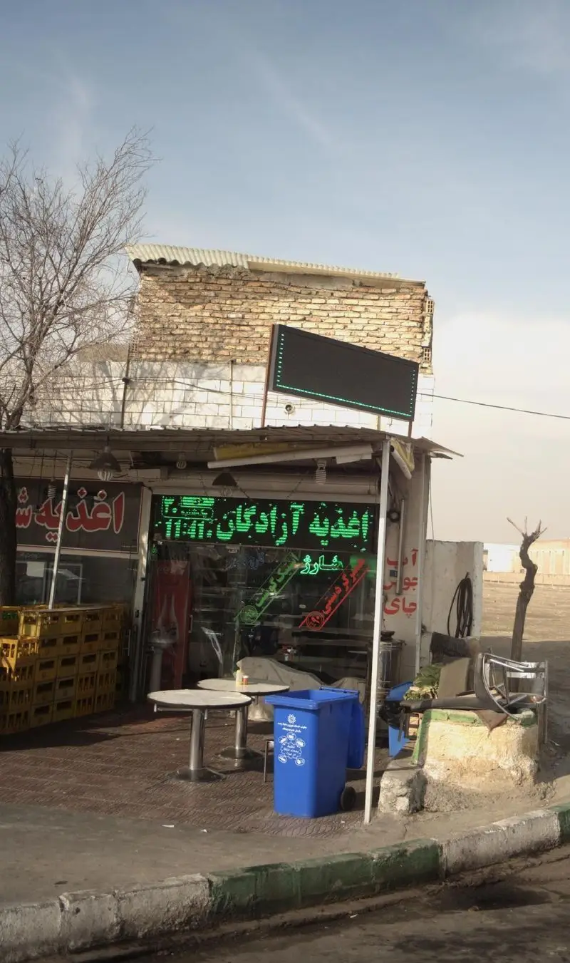 نمای بیرونی اغذیه آزادگان با تابلو LED سبز، میز و سطل بازیافت در محیطی ساده و صمیمی، ارائه دهنده غذای آماده.