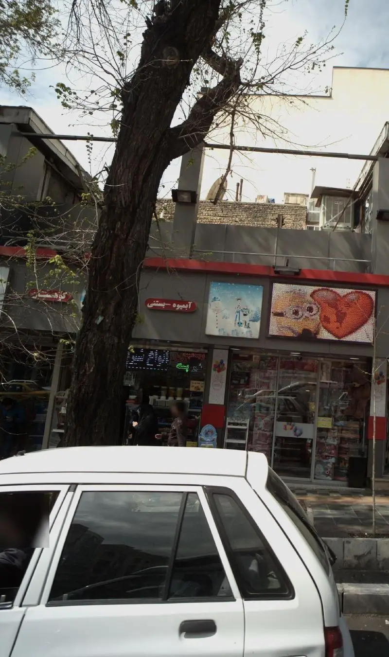 تصویر خیابانی از فروشگاه آبمیوه - بستنی کوهستان، با تابلوی روشن و مشتریان، مقابل درخت کهنسال و ماشین سفید.