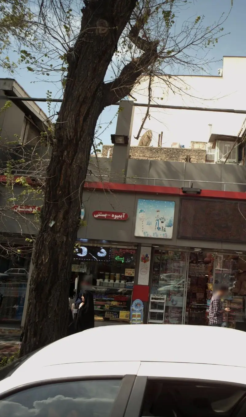 نمایی از فروشگاه آبمیوه و بستنی کوهستان در خیابان، شامل تابلوی مغازه، یک درخت بزرگ و اتومبیل پارک شده.