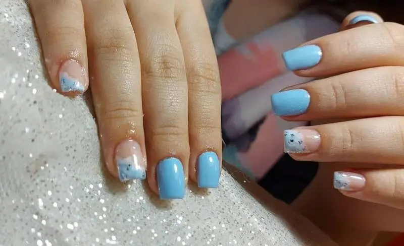 طراحی ناخن شیک و جدید با لاک آبی آسمانی ملایم و فرنچ اکلیلی نقرهای روی ناخنهای دست زنانه در سالن زیبایی تخصصی