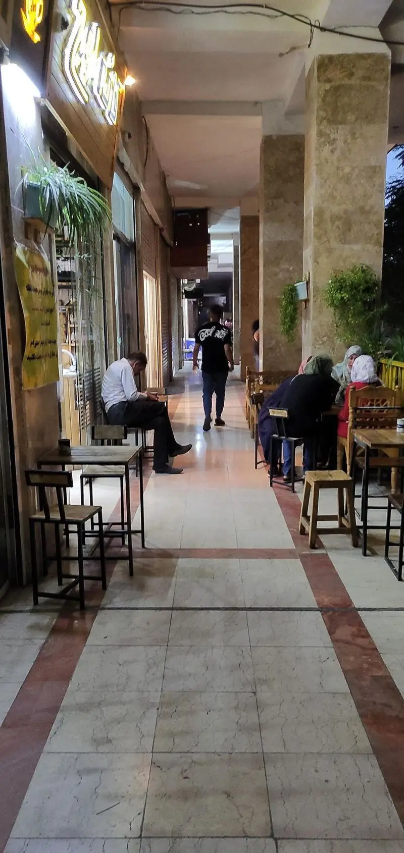 تصویری از نمای بیرونی کافه اوج در شب، با مشتریان نشسته بر روی میز و صندلی‌های چوبی در فضای باز راهروی سنگفرش.