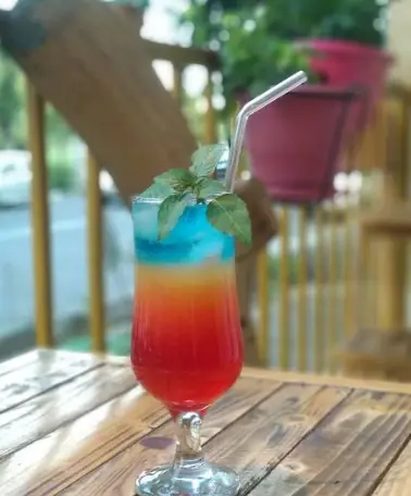 نوشیدنی لایه ای رنگارنگ و خنک با تزئین برگ نعنا و نی، سرو شده بر روی میز چوبی در کافه اوج. انتخابی جذاب از منوی ویژه.