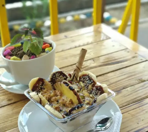 دسر خوشمزه کافه اوج: بستنی موز و شکلات با آجیل و ویفر. تصویری از محیط دنج و زیبای کافه اوج با گلدان روی میز.