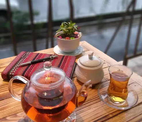 نوشیدن چای خوش‌رنگ در قوری شیشه‌ای و استکان زیبای کافه اوج، همراه با دفتر و قلم روی میز چوبی برای لحظات دنج و آرام.