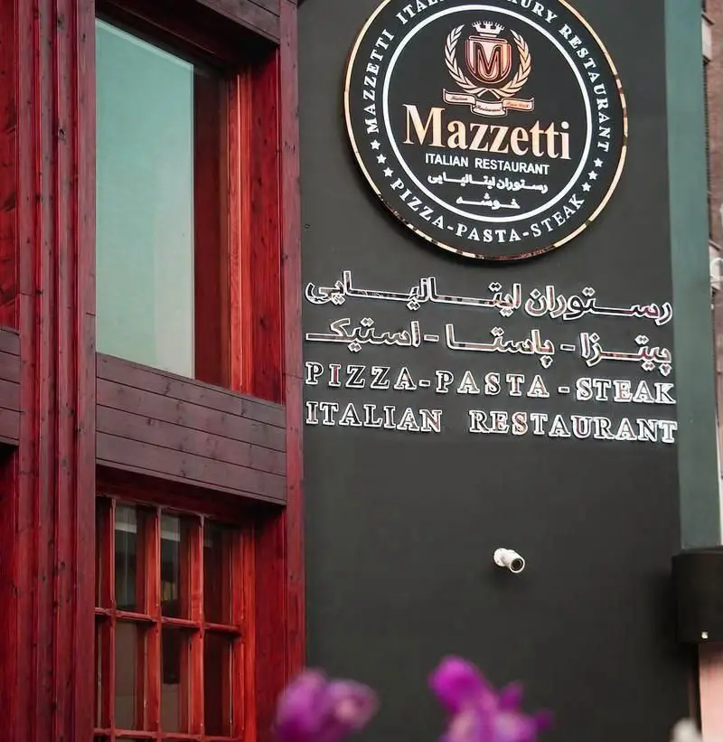 تابلوی مدرن رستوران ایتالیایی مازتی با شعار پیتزا، پاستا، استیک و نمای چوبی ساختمان در کنار آن.