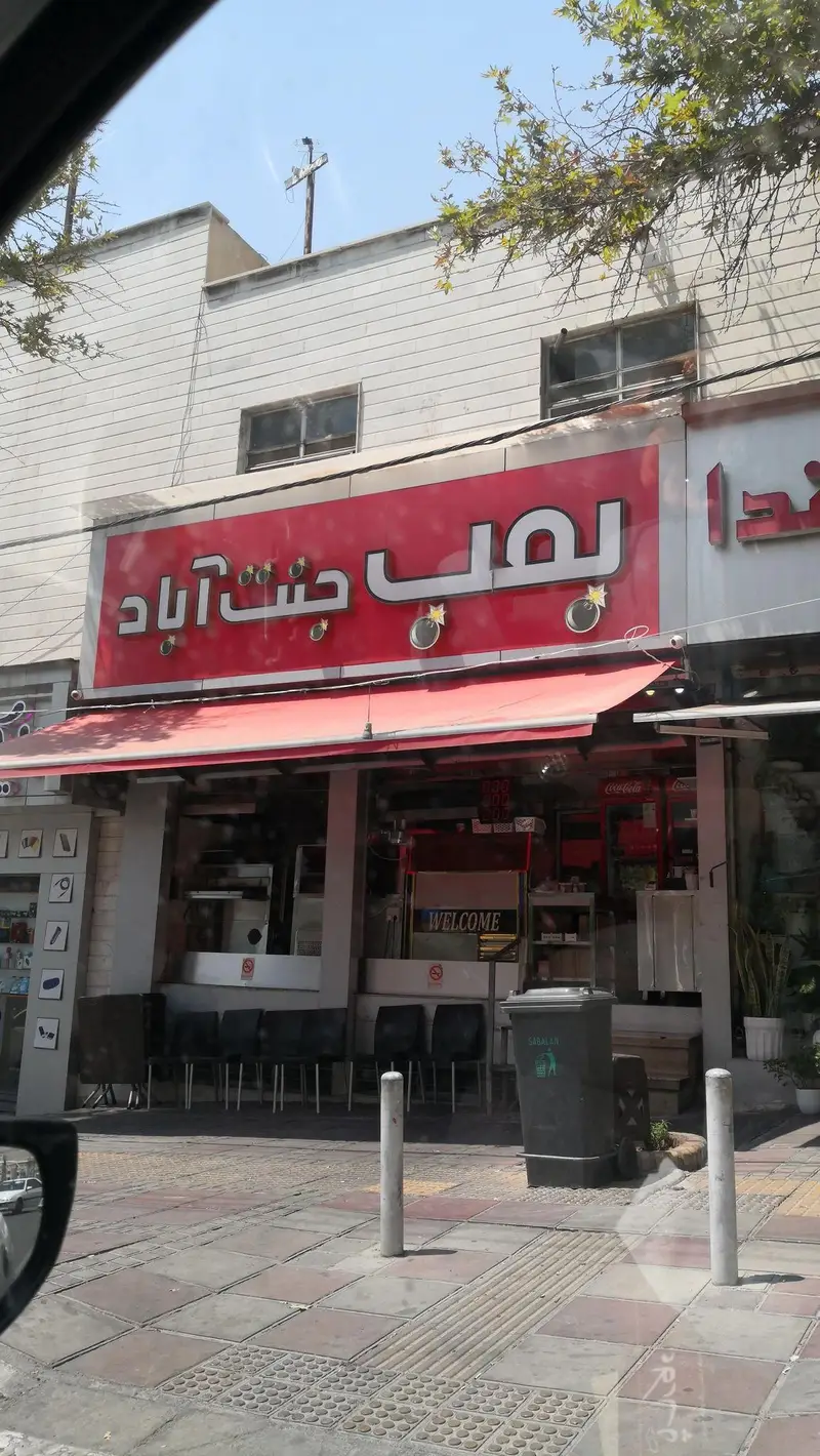 بمب جنت آباد: نمای بیرونی مغازه فست فود و ساندویچی معروف در منطقه جنت آباد تهران با تابلوی قرمز و چتر آفتابگیر.