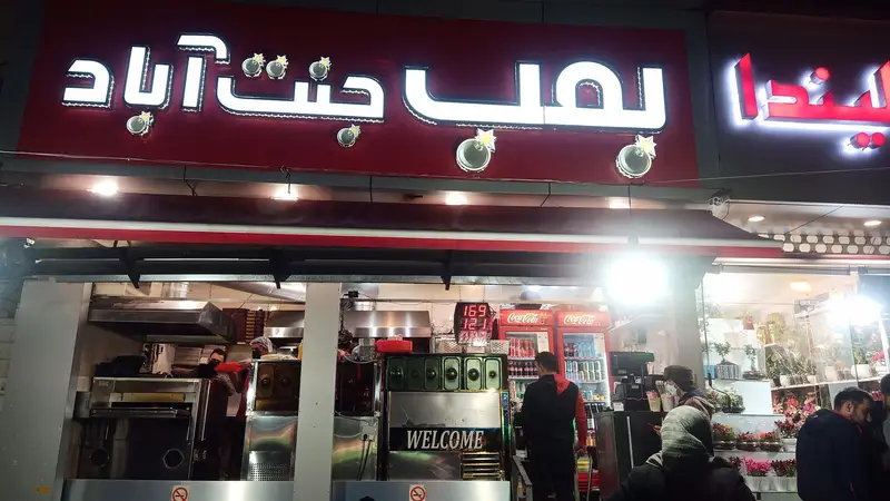 بمب جنت آباد: تصویر نمای جلوی رستوران فست فود بمب جنت آباد با تابلوی نئونی روشن، دکوراسیون شلوغ و مشتریان در فضای ورودی.