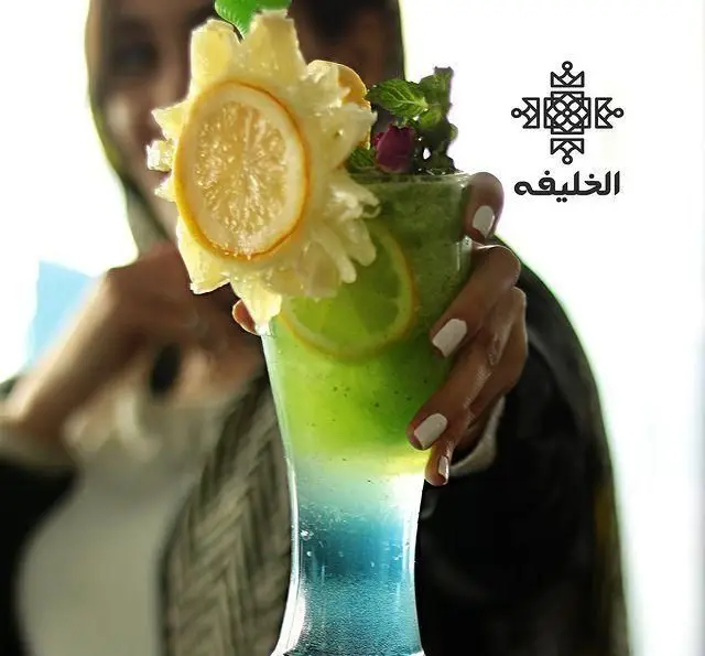 تصویری جذاب از یک نوشیدنی ترکیبی خنک و گوارا در کافه الخلیفه، با لایه‌های سبز و آبی و تزئینات هنرمندانه لیمو و گل. مناسب منوی رستوران الخلیفه.