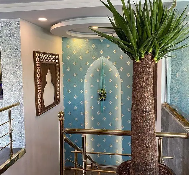 دکوراسیون شیک و جذاب رستوران و کافه الخلیفه، با دیوار آبی طرح‌دار، طاق زیبای سنتی، نرده‌های طلایی پله‌ها و گیاه یوکا بزرگ.