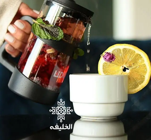تصویری از دست در حال سرو دمنوش میوه‌ای تازه و نعناع در لیوان شخصی، با تکه لیمو و غنچه گل رز در فنجان. نوشیدنی خاص کافه الخلیفه.