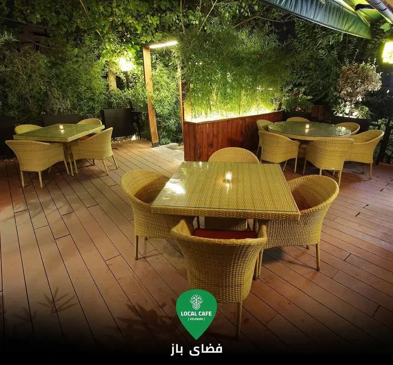 نمایی از فضای باز کافه لوکال ولنجک با چیدمان زیبای میز و صندلی‌های حصیری روی کفپوش چوبی و نورپردازی دلنشین گیاهان.