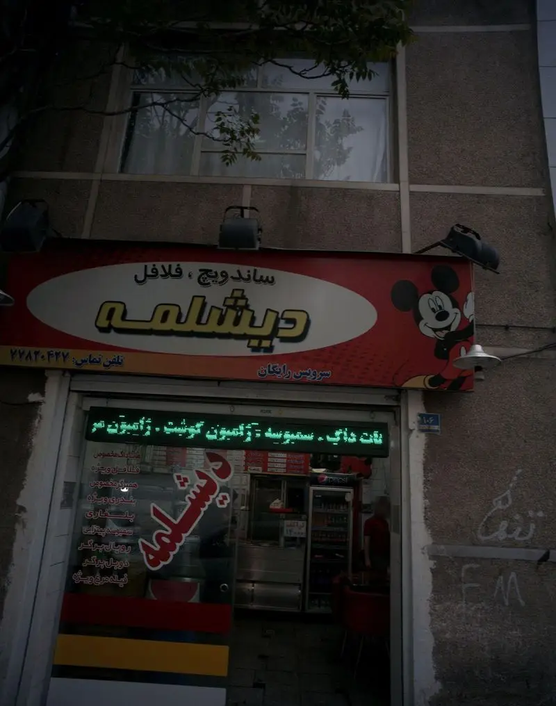 عکس از نمای بیرونی مغازه ساندویچ و فلافل دیشلمه با تابلوی بزرگ قرمز و میکی ماوس. انواع ساندویچ، فلافل، پیتزا، سمبوسه، نات داگ و ژامبون.