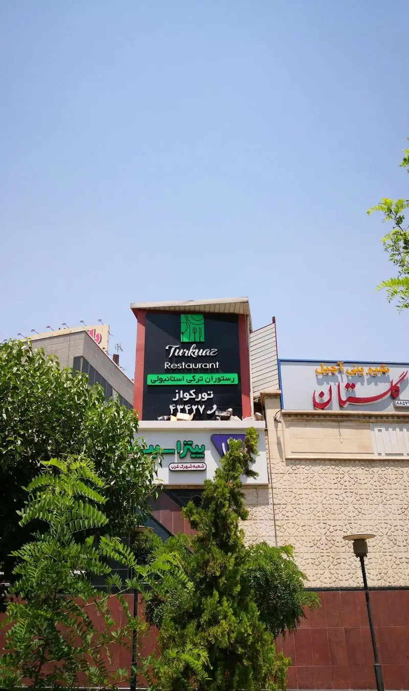 نمایی از تابلوی رستوران ترکی تورکواز با نام کامل «رستوران ترکی استانبولی تورکواز» در یک روز آفتابی با آسمان آبی و درختان سرسبز.
