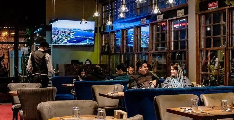 فضای داخلی دنج رستوران ترکی تورکواز با دکوراسیون مدرن، مشتریان خوشحال، گارسون فعال و نمایش مناظر شهری. تجربه غذایی اصیل ترکی.