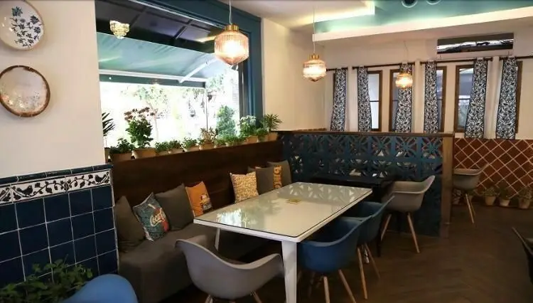 فضای دلنشین کافی شاپ ایل با مبلمان راحتی، میز و صندلی شیک، کاشی‌کاری آبی، گلدان‌ها، و نور طبیعی کنار پنجره.