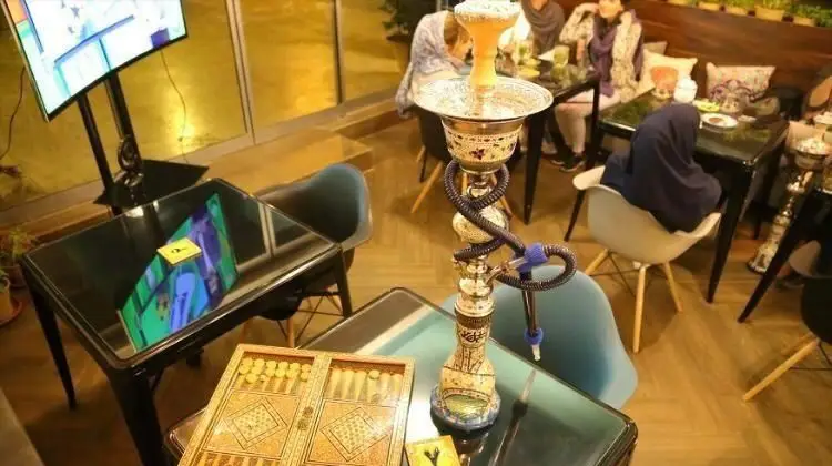 نمای داخلی کافی شاپ ایل با مشتریان، قلیان و تخته نرد. فضایی دلنشین برای دورهمی و استراحت در کافه با تلویزیون بزرگ.