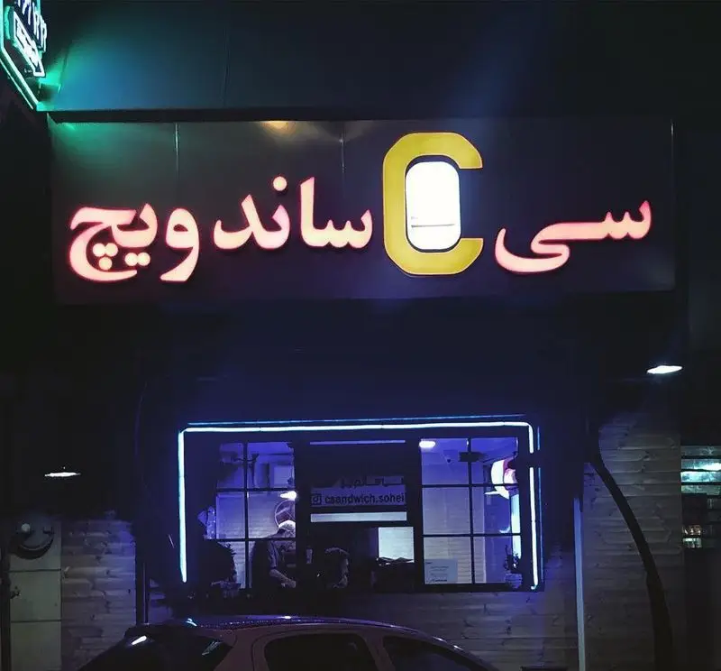 نمایی از تابلوی نئونی درخشان سی ساندویچ در شب، با ویترین روشن و نور آبی فست فود. بهترین گزینه برای ساندویچ های لذیذ و پرطرفدار.