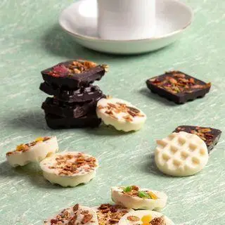 انواع شکلات‌های دست‌ساز کافه قنادی کوک، شامل شکلات تلخ و سفید با تزئینات لوکس، در کنار فنجان قهوه، مناسب پذیرایی و دسر.