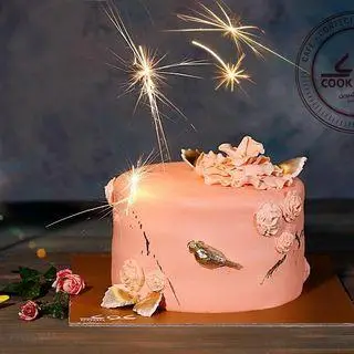 کیک تولد صورتی شیک با تزیین پرنده و گل‌های برجسته طلایی، همراه با فشفشه، از کافه قنادی کوک برای لحظات شیرین شما.