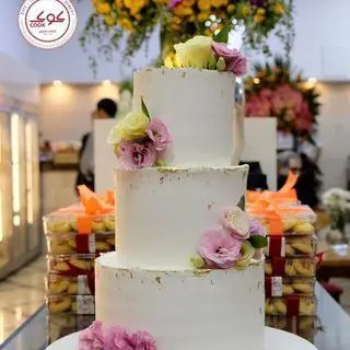 کیک عروسی سه طبقه سفید و لوکس کافه قنادی کوک با تزیین گل طبیعی صورتی و زرد، مناسب برای سفارش کیک عروسی و تولد.