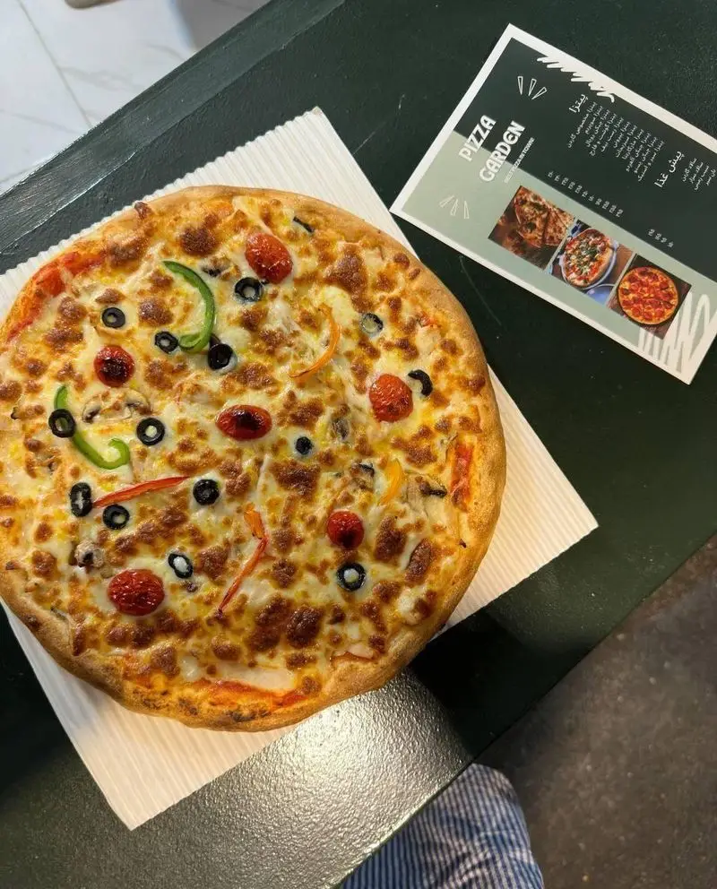 پیتزای تازه و لذیذ پیتزا گاردن با گوجه گیلاسی، زیتون سیاه، قارچ و فلفل دلمه‌ای رنگارنگ، آماده سرو روی میز، در کنار منوی سبز.