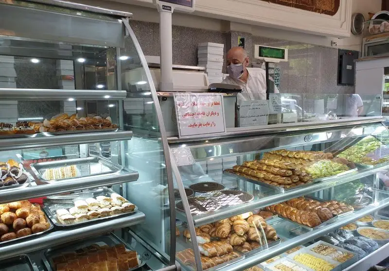 تصویر داخلی قنادی و شیرینی فروشی کسری. ویترین‌ها پر از انواع شیرینی‌های سنتی و مدرن، تر و خشک. فروشنده با ماسک.