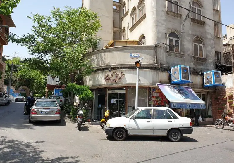 مغازه شیرینی کسری در یک خیابان آفتابی شهر، با خودروهای پارک شده، ساختمان قدیمی و درختان سرسبز، نمایی از زندگی شهری