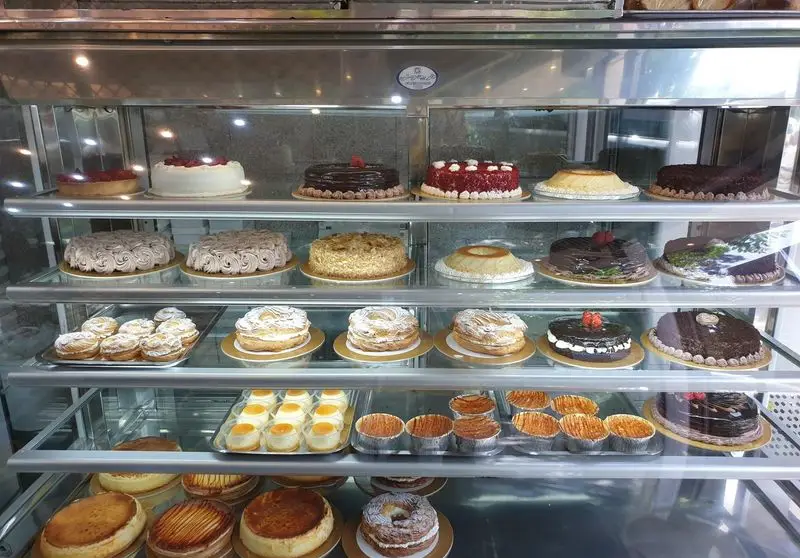 ویترین جذاب شیرینی کسری با ردیف‌های پر از کیک‌های تازه، شامل کیک شکلاتی، ردولوت، تارت میوه و دسرهای مجلل و شیرینی‌های متنوع و لذیذ.