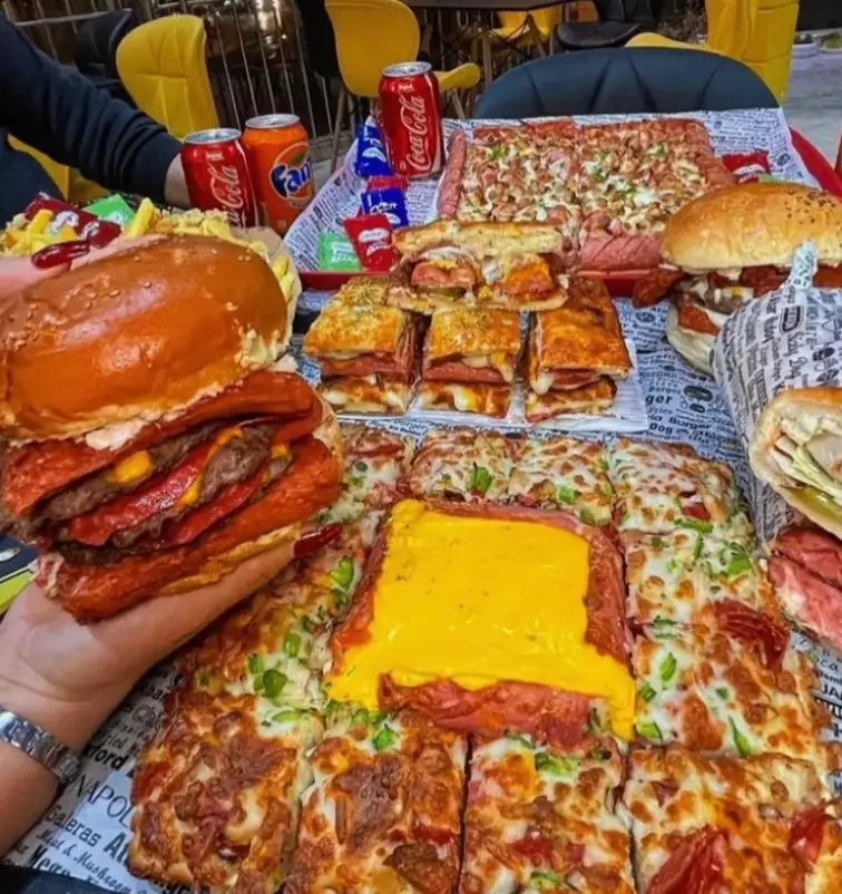 میز پر از غذاهای فست فود عطاویچ فودکورت خلیج فارس، با برگر غول‌پیکر، پیتزاهای متنوع، ساندویچ‌ها، سیب‌زمینی و نوشابه.