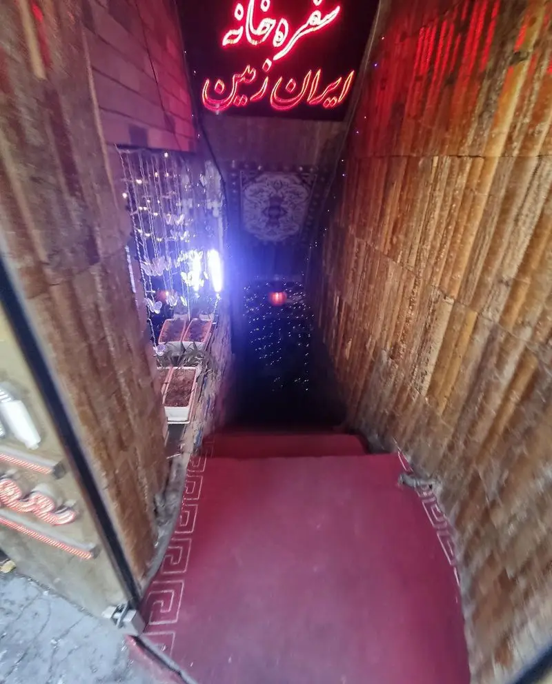 ورودی دنج و زیبای سفره خانه سنتی ایران زمین، با پله‌های پوشیده از فرش قرمز، دیوارهای چوبی و نورپردازی دلنشین و دعوت‌کننده.