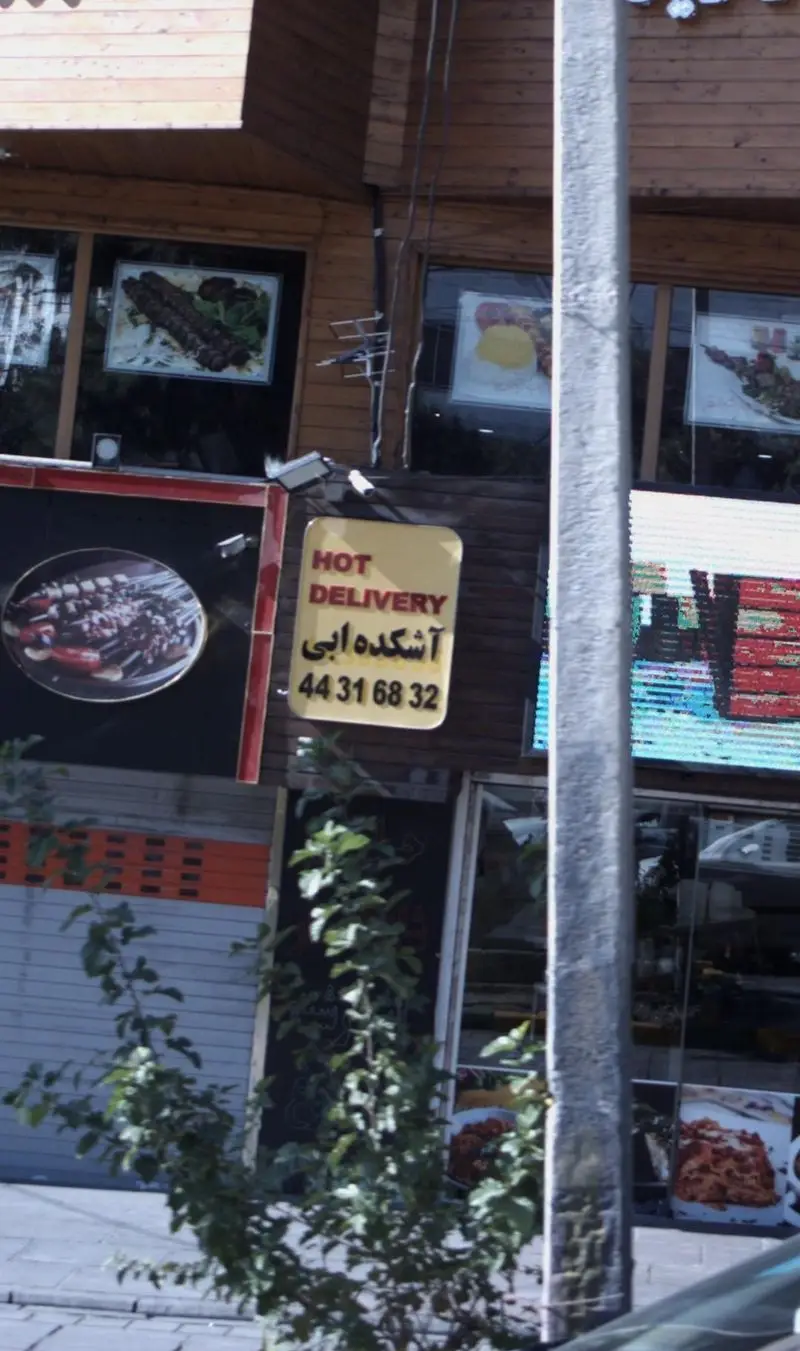 نمای مغازه آشکده ابی با سردر چوبی و تابلوی "HOT DELIVERY" و شماره تماس 44316832. ویترین آن شامل تصاویری از انواع کباب و آش ایرانی برای سفارش.