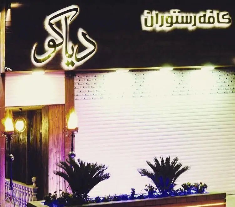 تصویر شب از کافه رستوران دیاکو با تابلوی نئون و ورودی روشن. نمای بیرونی جذاب با گلدان‌های گل و معماری چوبی، محیطی دلنشین برای پذیرایی.
