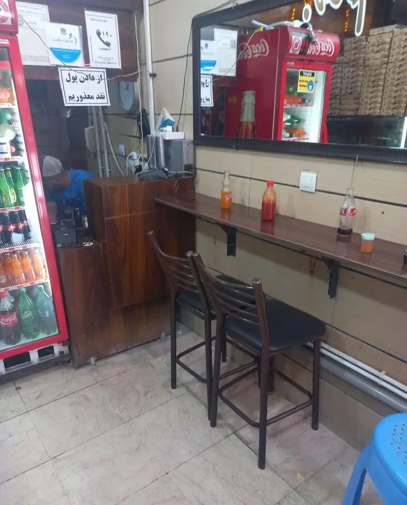محیط داخلی فست فود بهنام با میز و صندلی، یخچال نوشیدنی کوکاکولا و پیشخوان آماده برای سفارش غذا و نوشیدنی.