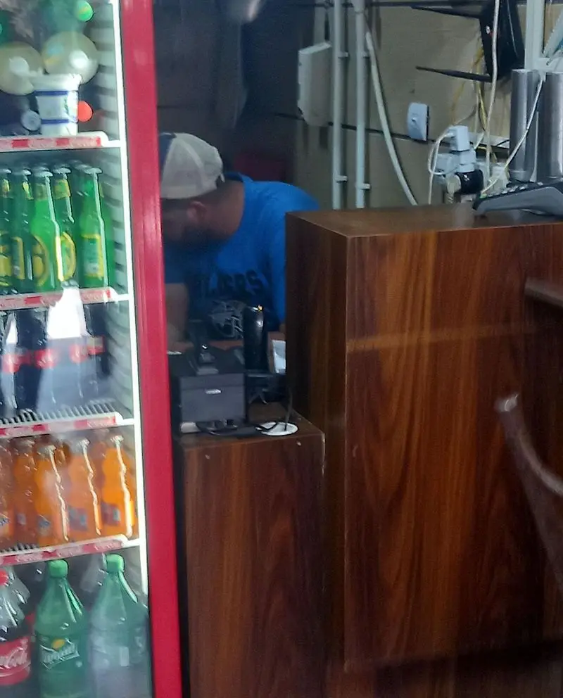 کارمند فست فود بهنام با کلاه و تی‌شرت آبی، مشغول کار پشت پیشخوان چوبی، در کنار یخچال مملو از انواع نوشیدنی‌های خنک و بطری.